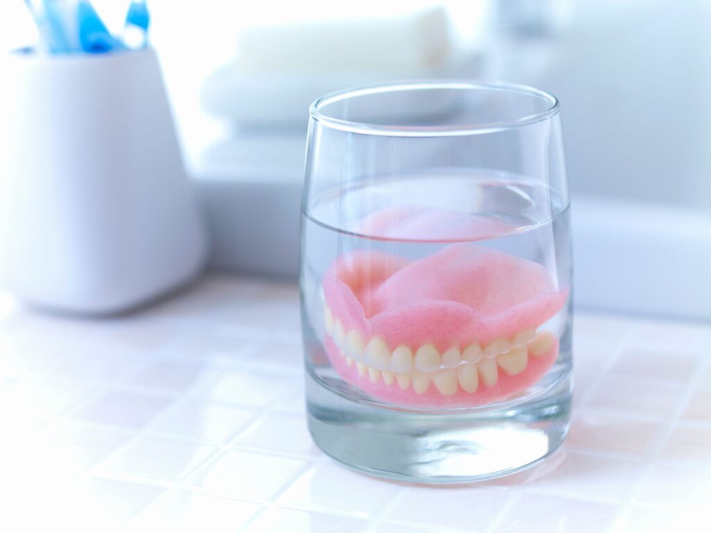 Full set of dentures in a glass of clear liquid on tiled countertop